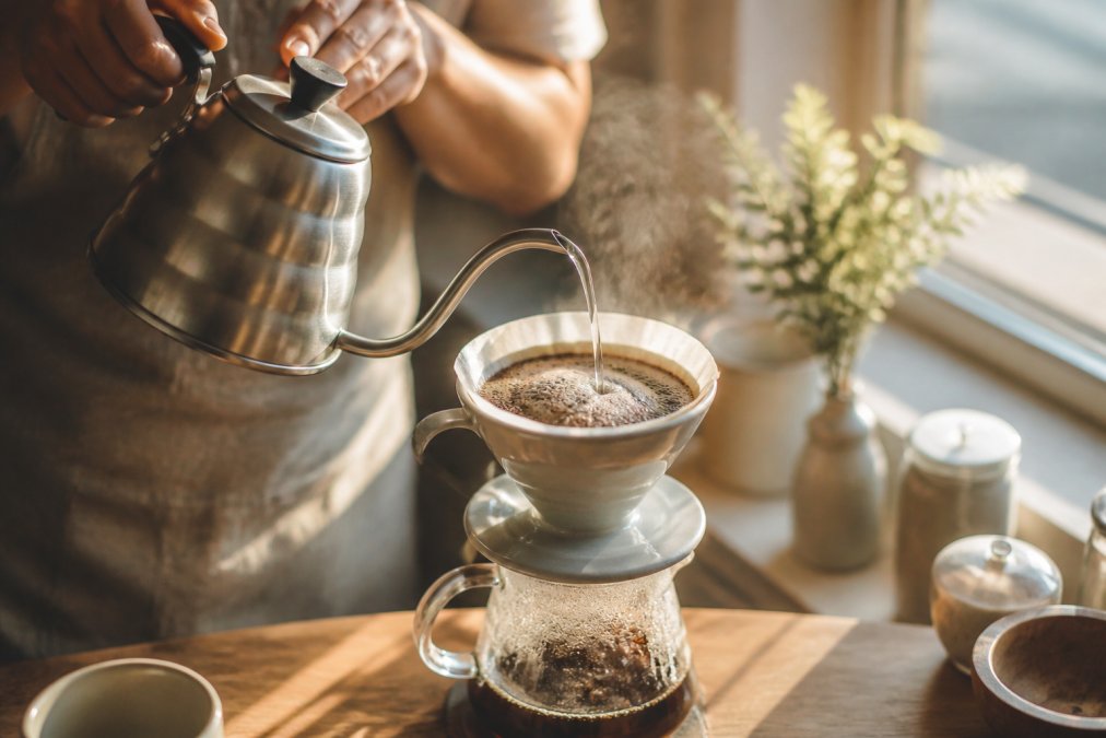 Close-up of hands pouring hot water in slow circular motions over freshly ground coffee in a ceramic pour-over dripper, steam rising visibly, warm morning sunlight streaming through a kitchen window