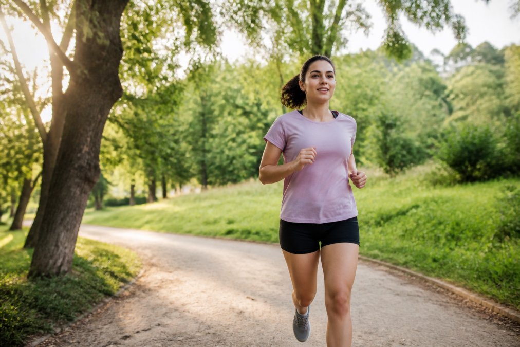 Person jogging outdoors in morning light through a park with trees, appearing energized and focused, natural sunlight filtering through branches