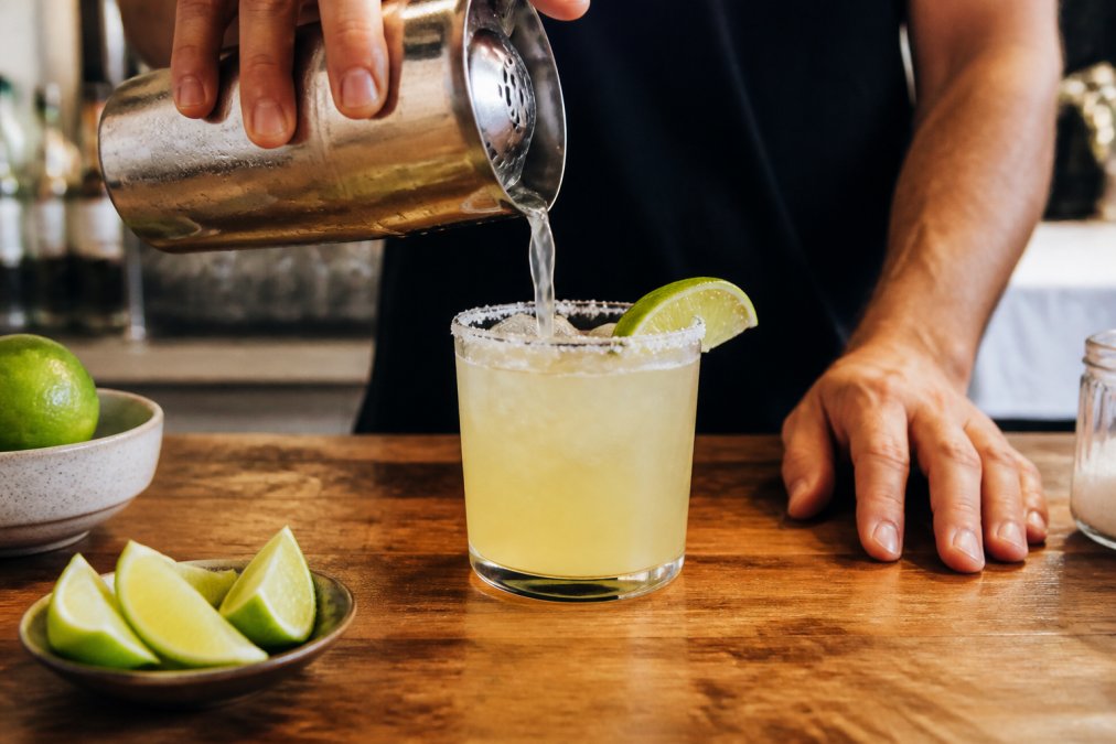 Hands pouring a fresh tequila cocktail into a chilled glass with a lime wedge on a wooden bar top for simple recipes.