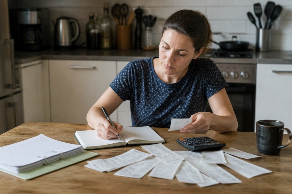 Person at kitchen table with notebook, receipts, and calculator, reviewing household budget and expenses with organized papers spread out