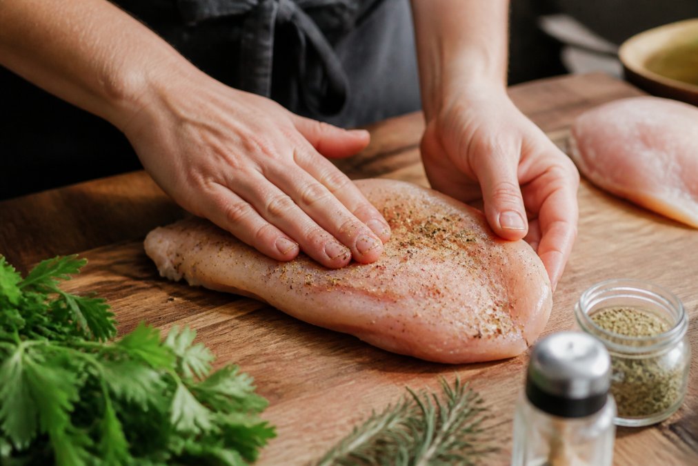 Close-up of hands rubbing seasoning blend onto raw chicken breast on a wooden cutting board with fresh herbs and spice containers nearby
