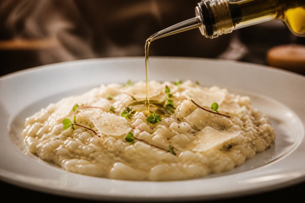 Close-up of creamy risotto with truffle oil being drizzled over the top, steam rising, garnished with fresh parmesan and microgreens on white plate with warm lighting