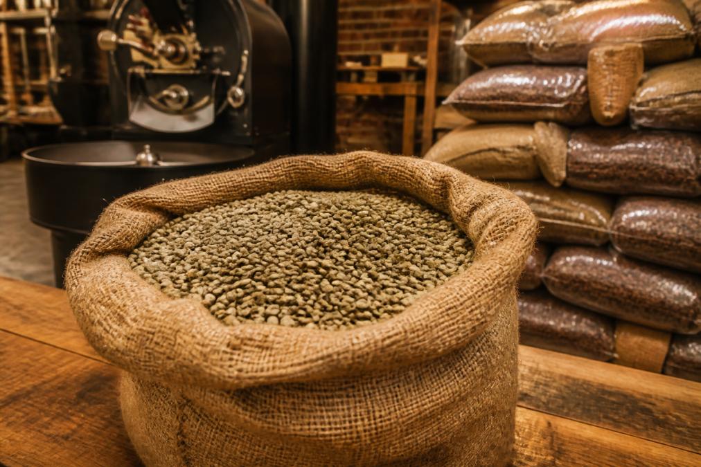 A burlap sack filled with green coffee beans sits on a wooden table in a roastery, with a roasting machine and stacked bags of roasted beans softly blurred in the background