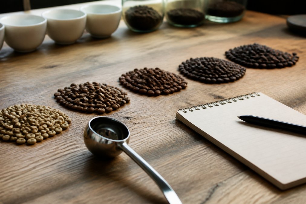 Close-up of specialty coffee beans from different origins arranged in small piles showing color variation from light to dark roasts, with a coffee cupping spoon and tasting notes card on a wooden surface with natural window lighting
