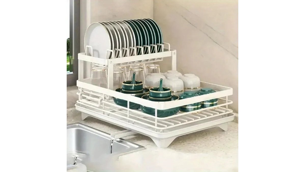 Stainless steel compact dish rack with angled drip tray positioned next to a modern chrome faucet on white countertop in minimalist apartment kitchen with natural morning light