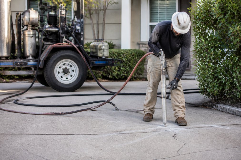 A technician in safety gear using injection equipment on a residential driveway, with hoses and a small rig visible, demonstrating non-invasive soil stabilization work