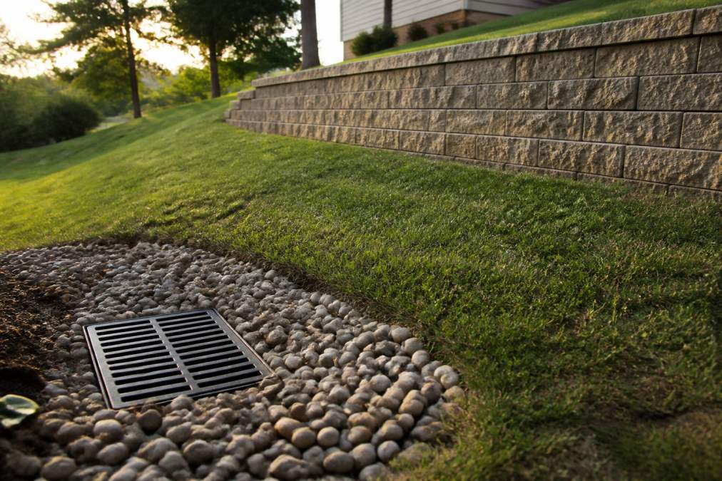 A Metro Detroit backyard with a sloped lawn, a modular block retaining wall in the background, and a visible French drain grate in the foreground during late afternoon light