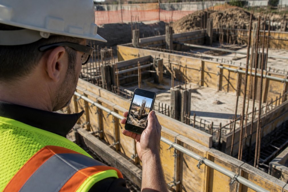 A field inspector wearing a hard hat and high-visibility safety vest holding an Android smartphone on an active construction site, reviewing a geotagged photo with typed notes visible on the screen, afternoon sunlight casting shadows across concrete formwork