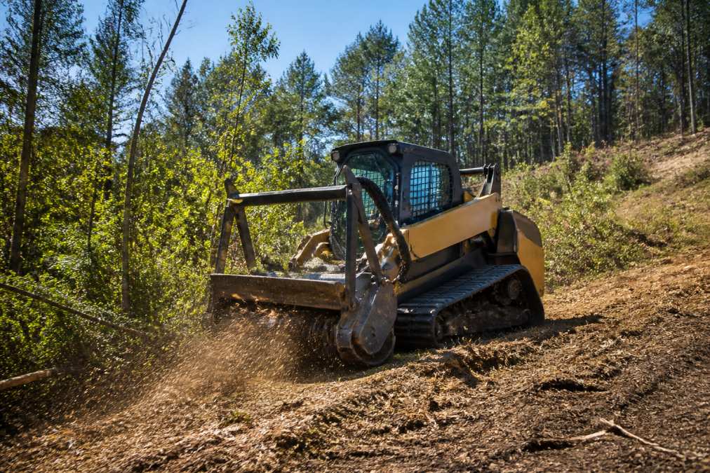 Forestry mulching equipment actively grinding dense brush and small trees on sloped Central Virginia property, with mulch being ejected and topsoil visible underneath, afternoon sunlight