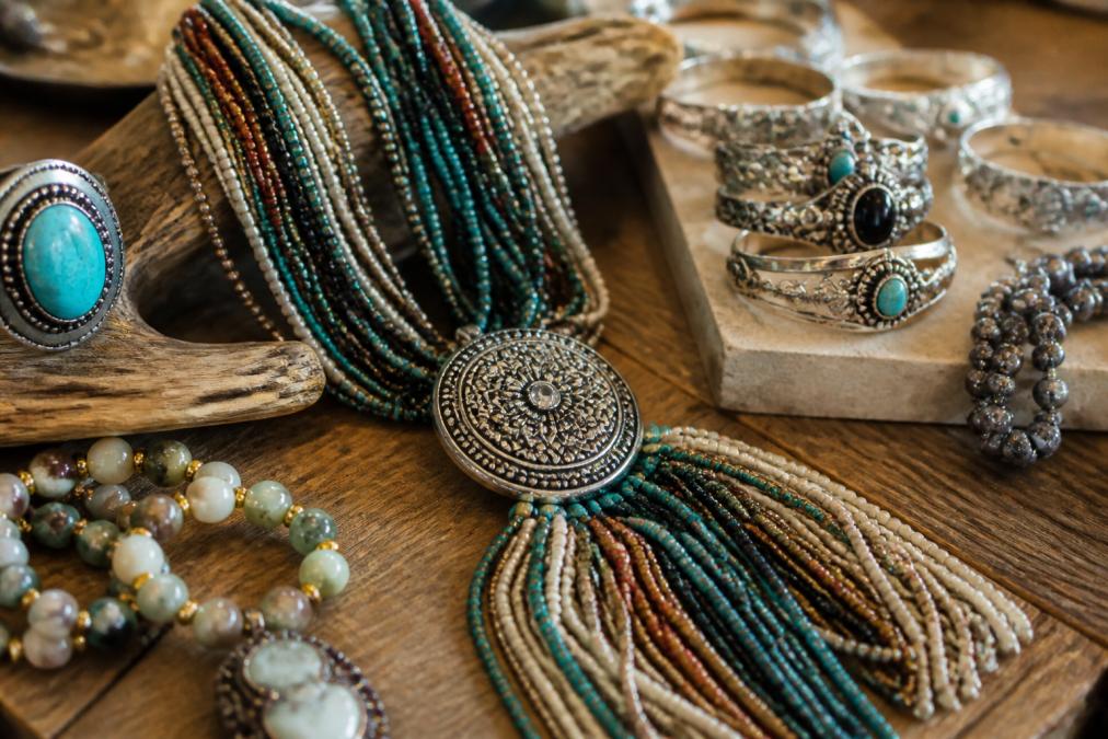 Close-up of high-quality statement jewelry and accessories displayed on natural wood surface, showing intricate beading, metalwork detail, and craftsmanship in warm natural lighting