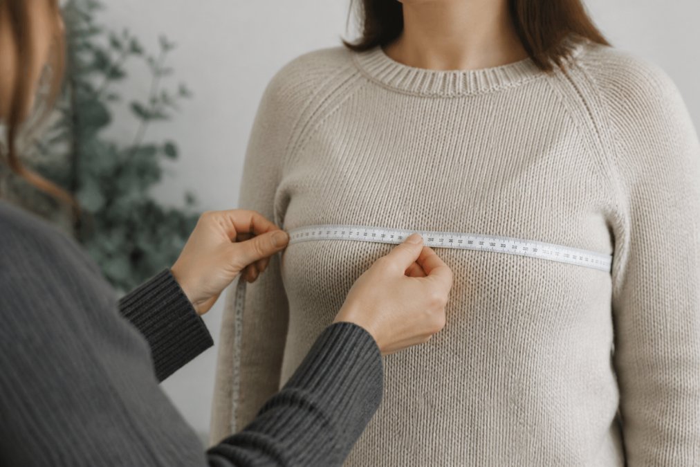 Close-up of hands measuring a person's bust circumference with a flexible tape measure positioned parallel to the floor, showing proper measurement technique with natural studio lighting