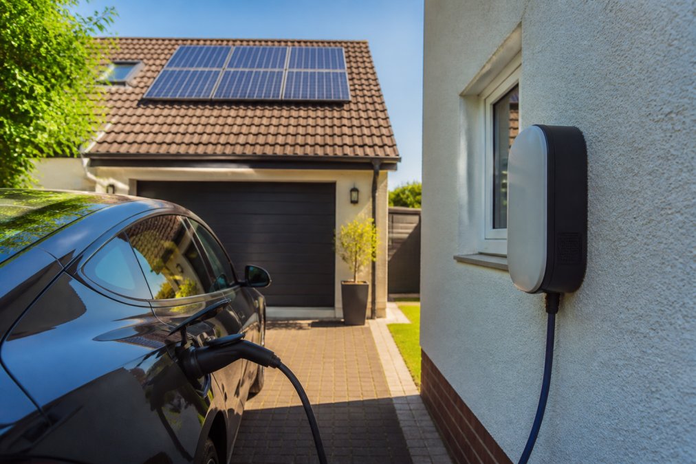 Electric vehicle charging at a home wall-mounted charger during bright daylight with solar panels visible on the roof and clear sky
