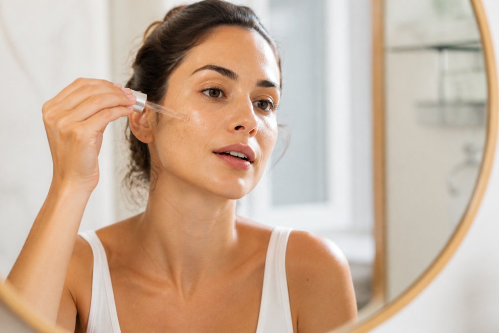 Close-up of a woman's hand gently applying a clear serum dropper to her cheek, soft natural lighting, clean bright bathroom background
