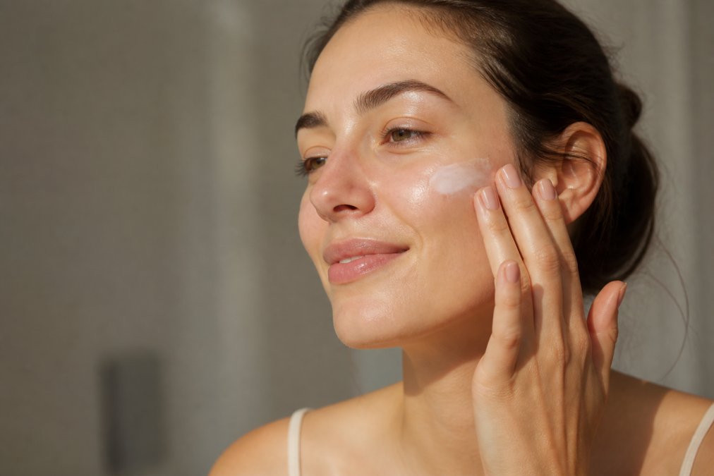 Close-up of a woman's hand applying a lightweight moisturizer to her face with gentle upward strokes, natural soft lighting illuminating calm, hydrated skin with a healthy glow