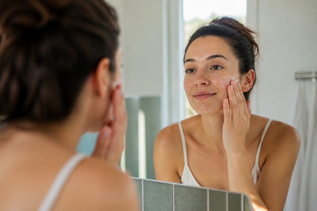 Woman with calm, glowing skin applying a gentle moisturizer to her face in a bright, minimalist bathroom with soft natural lighting