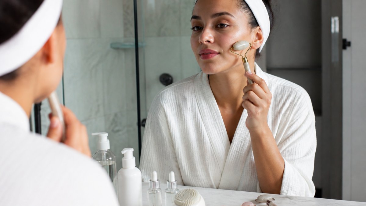Woman using a facial roller on her face during a morning skincare routine, with cleansing products visible on a bathroom counter