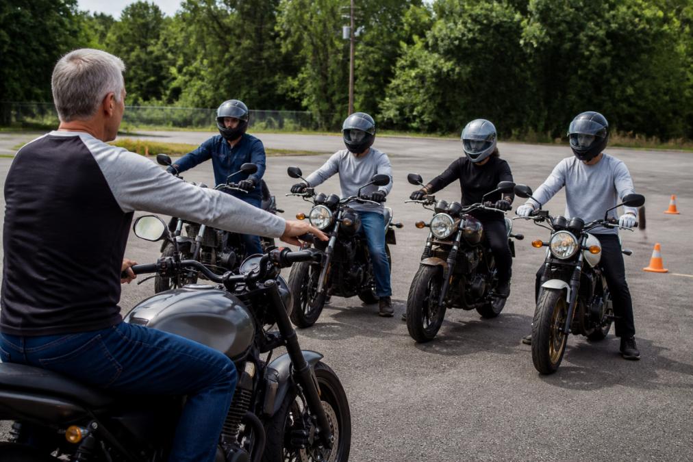 Certified motorcycle instructor demonstrating proper riding posture and hand positioning to a small group of students during a hands-on training session on a closed course