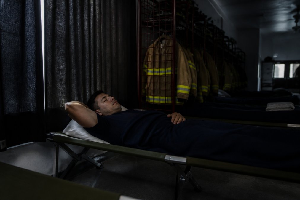A firefighter in a dark, quiet bunkroom at the fire station lying on a cot with blackout curtains visible, demonstrating an optimized sleep environment