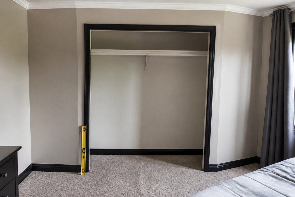 Bedroom wardrobe opening showing visible uneven walls, dark skirting boards at the base, carpeted floor with a spirit level positioned against the frame to demonstrate out-of-square conditions and measurement challenges
