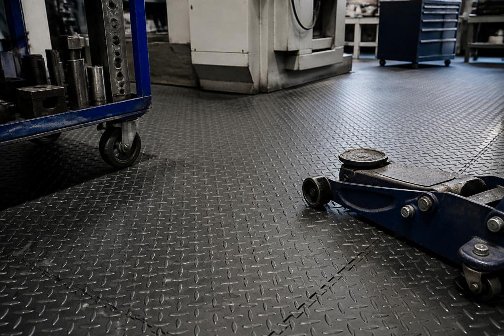 Tæt billede af slidstærke Fortelock PVC-fliser installeret i værkstedsmiljø med tunge metalværktøjer og industriudstyr på gulvet, viser overfladestrukturen og holdbarhed under belastning