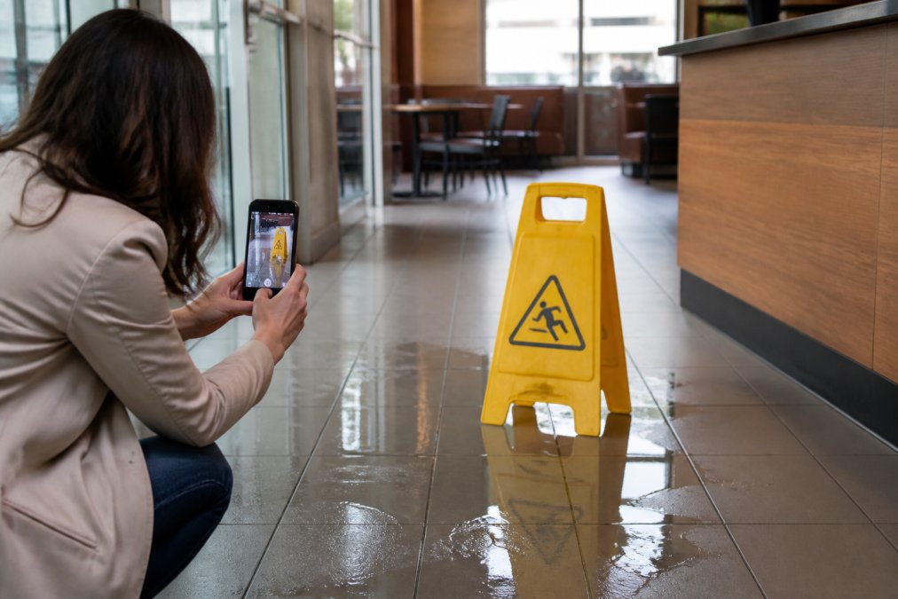 Person taking photos of a wet floor hazard with their smartphone, documenting the accident scene with visible wet surface and caution signs in a commercial setting