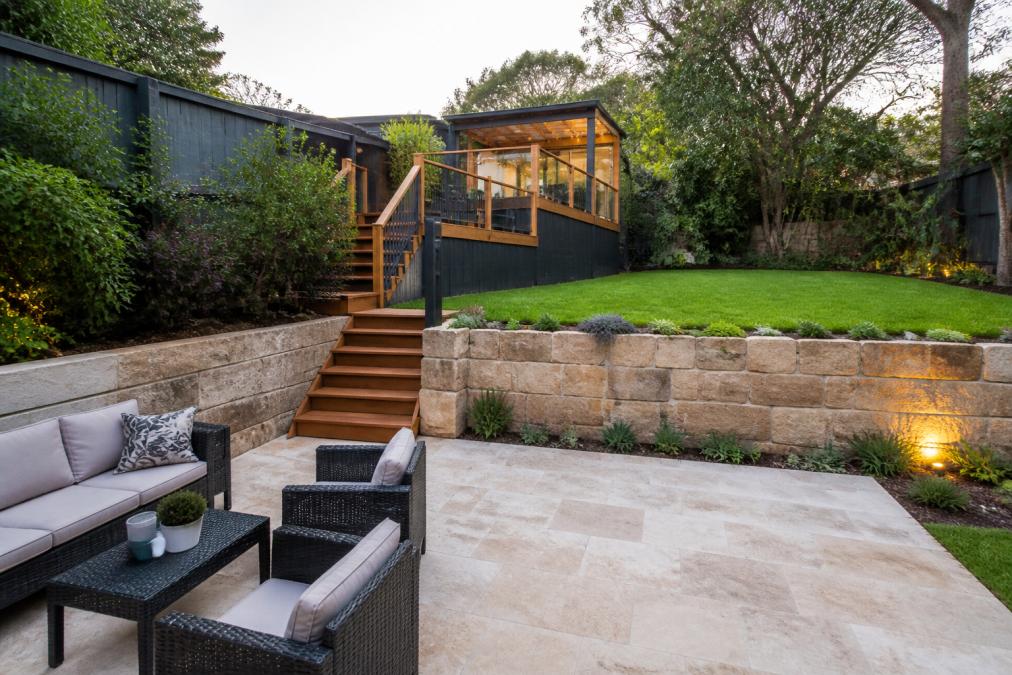 A completed multi-level backyard on a slope in Sydney, featuring a sandstone retaining wall, timber deck, and lush lawn, with outdoor furniture on a paved patio area