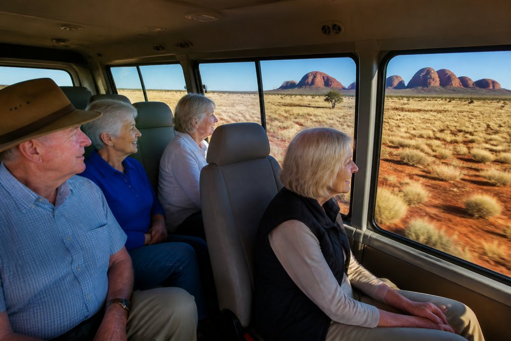 Small group of mature travellers seated comfortably in a purpose-designed 4WD vehicle with wide windows, smiling while observing a vast outback landscape of red earth, sparse spinifex grass, and distant rocky formations under clear blue sky