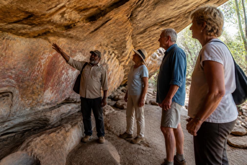 Aboriginal guide pointing out details and stories on ancient ochre rock art to small group of mature tourists standing in natural rock shelter with warm natural light filtering through opening, surrounded by weathered stone walls