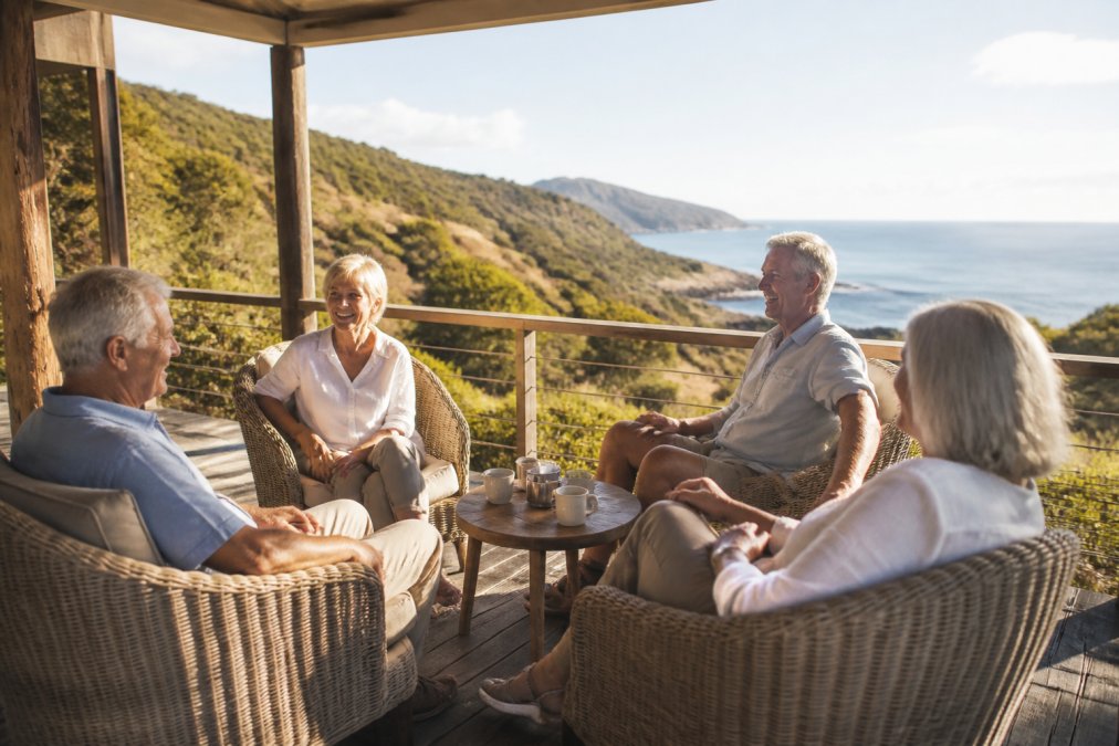 Group of mature travellers relaxing in comfortable woven chairs on a shaded veranda overlooking a coastal landscape, smiling and engaged in conversation, with soft afternoon light creating a peaceful atmosphere