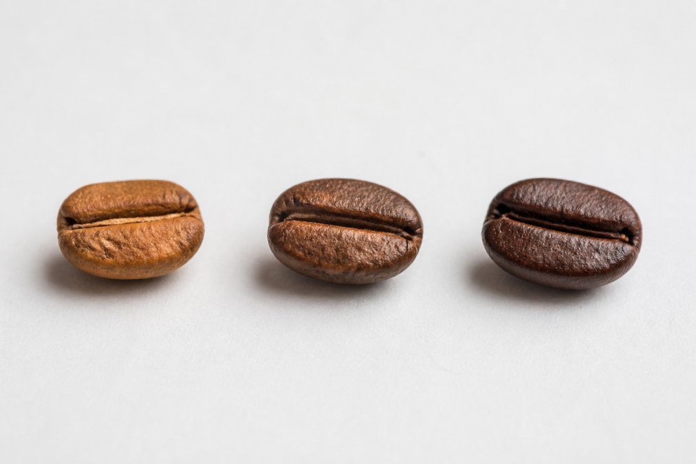 Close-up of three individual coffee beans showing distinct color progression from light brown to medium brown to dark brown, arranged in a line on a neutral white surface with natural window lighting emphasizing the color variations