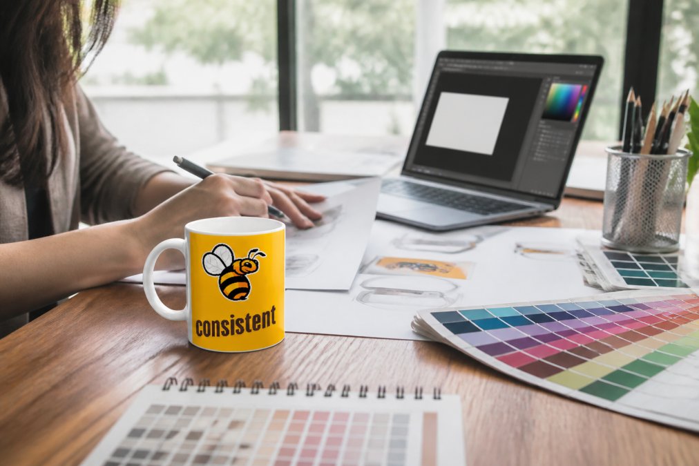 Flat lay of a custom ceramic mug with personalized logo design on a wooden desk, surrounded by design templates, color swatches, and a laptop displaying design software interface
