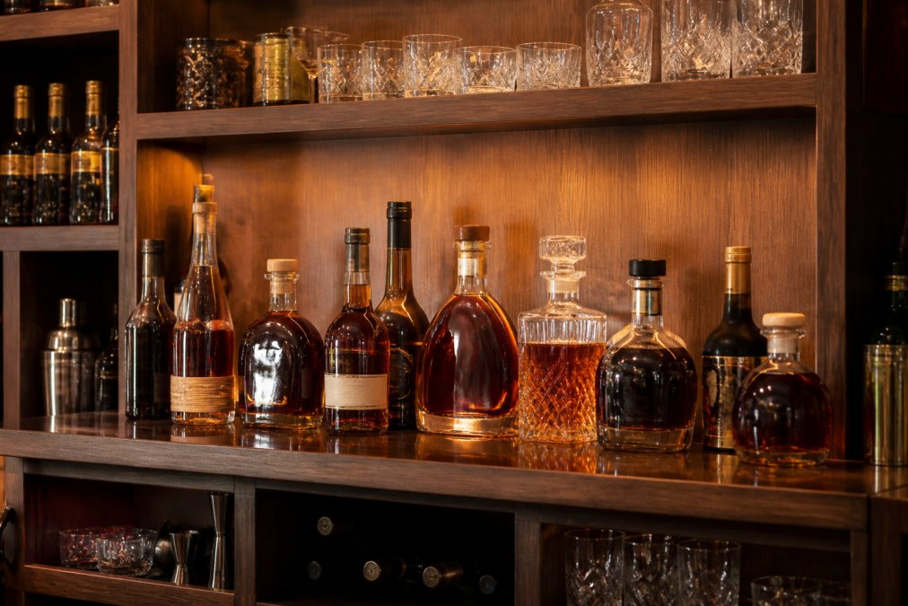 Premium rum bottles arranged on a wooden home bar shelf with crystal glassware, amber lighting highlighting the bottles, showing a curated collection of craft spirits ready for sipping or cocktail preparation