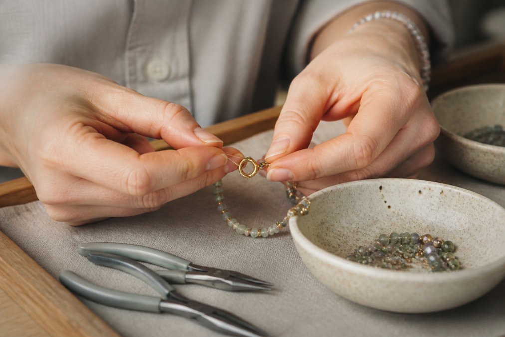 Close-up of hands carefully hand-stringing gemstone beads with gold spacers and clasp, showing meticulous detail work in natural light