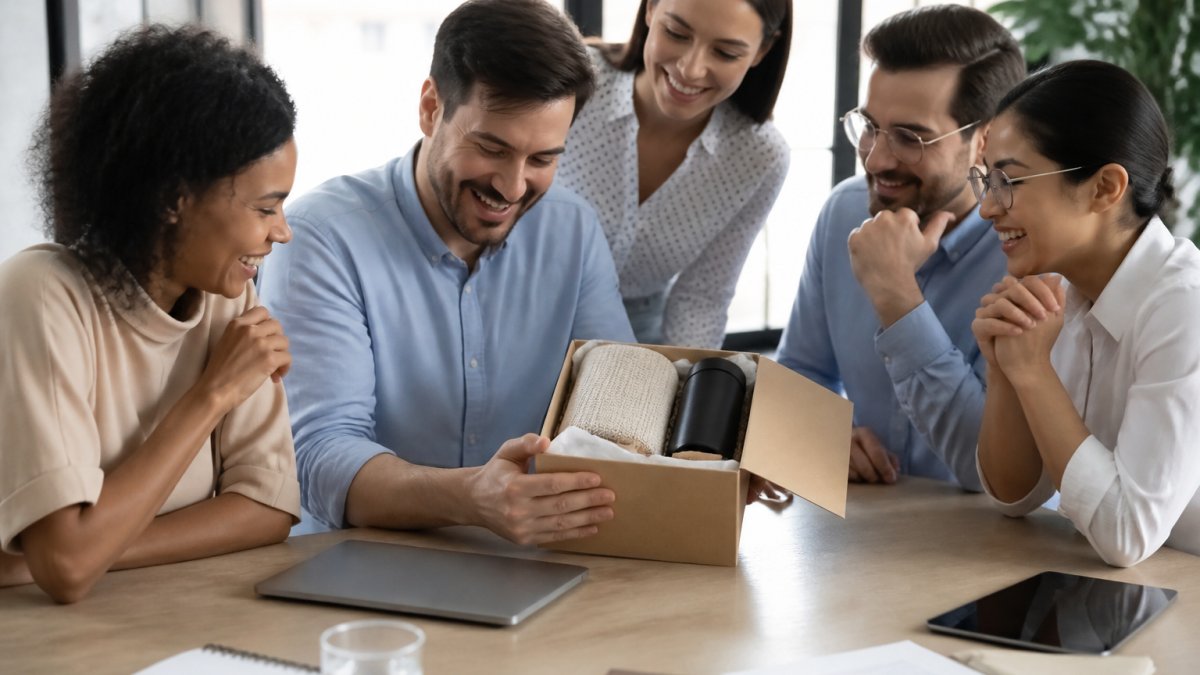 Small team of employees gathered around a conference table, opening a personalized gift box together, smiling with genuine delight, natural office lighting from large windows