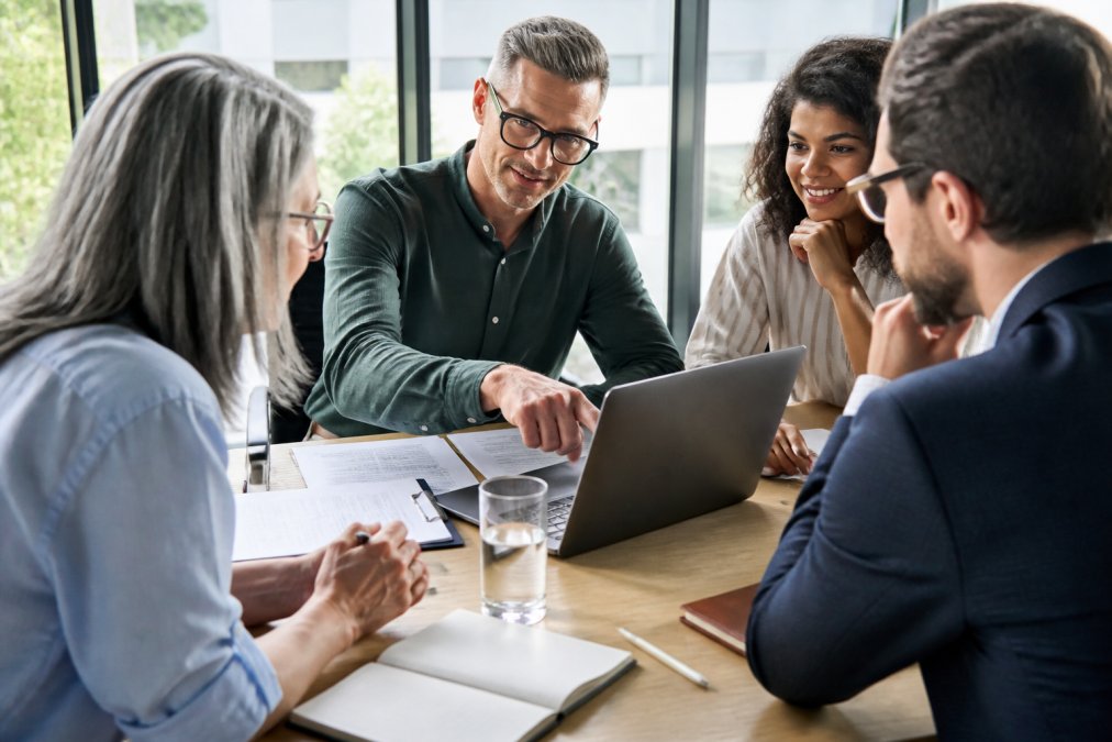 Marketing team members collaborating around a table, pointing at performance metrics displayed on a laptop screen in a bright office setting