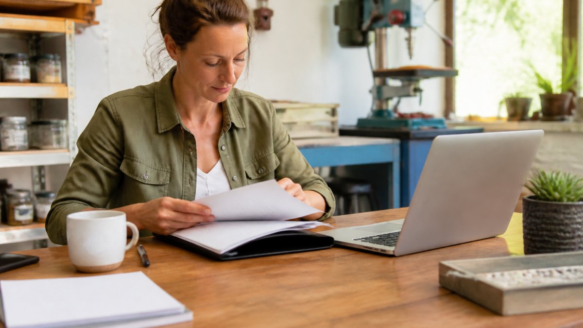 A small business owner at a tidy wooden desk reviewing a folder of new-hire paperwork beside a laptop showing a payroll dashboard, coffee mug and pen within reach, soft morning window light