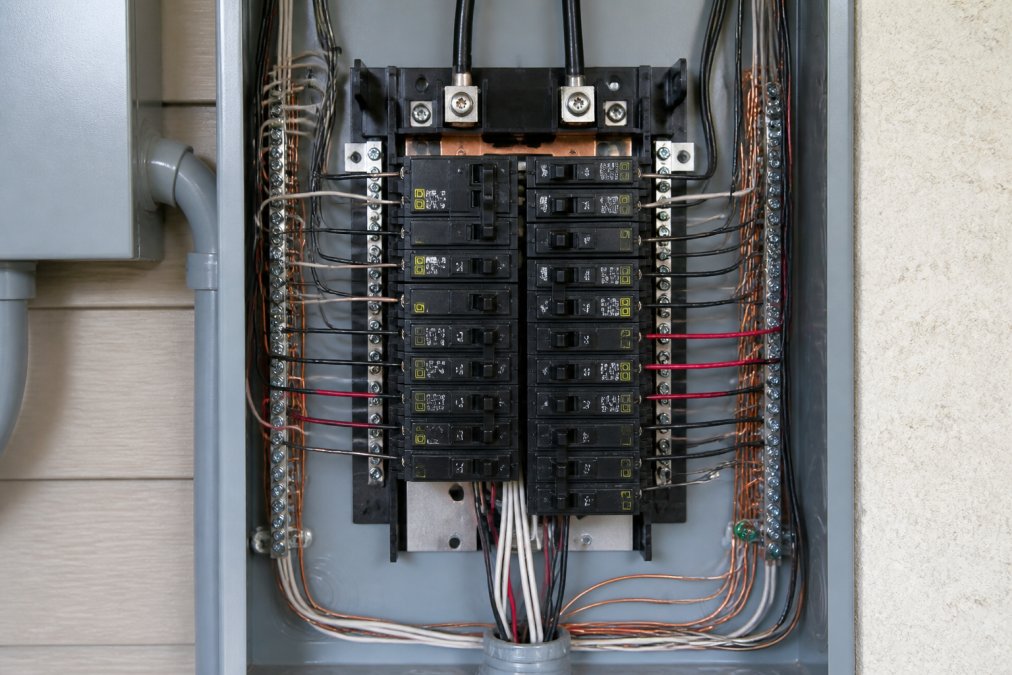 Close-up of an open residential electrical panel showing multiple circuit breakers arranged in rows, clearly labeled breaker switches, copper bus bars, and color-coded wiring connections with proper grounding terminals visible