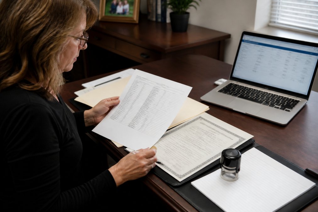 Family member sitting at a desk with legal documents, death certificate, notary stamp visible, and a laptop showing account information, reviewing paperwork with focused attention in natural office lighting