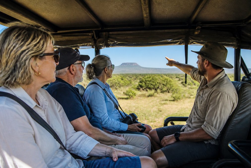 A small group of mature travellers sitting together inside a purpose-designed 4WD vehicle, engaged in conversation with a guide who is pointing toward a distant landscape feature, showing relaxed interaction and personal attention