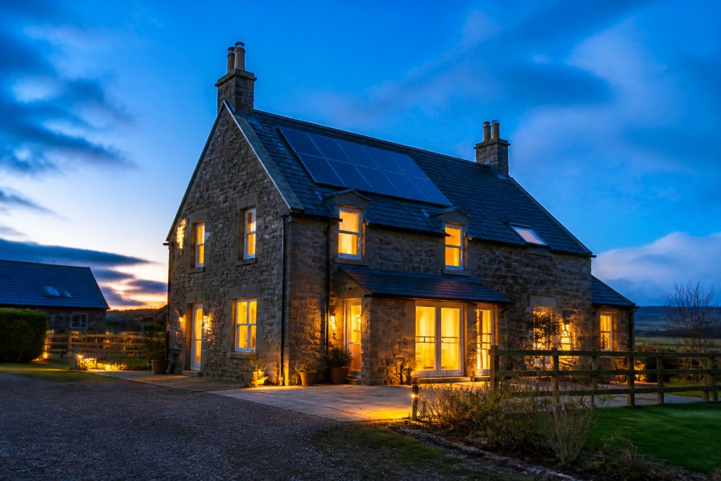 A modern Scottish stone home at dusk with solar panels on the roof, warm light glowing from the windows against a blue twilight sky