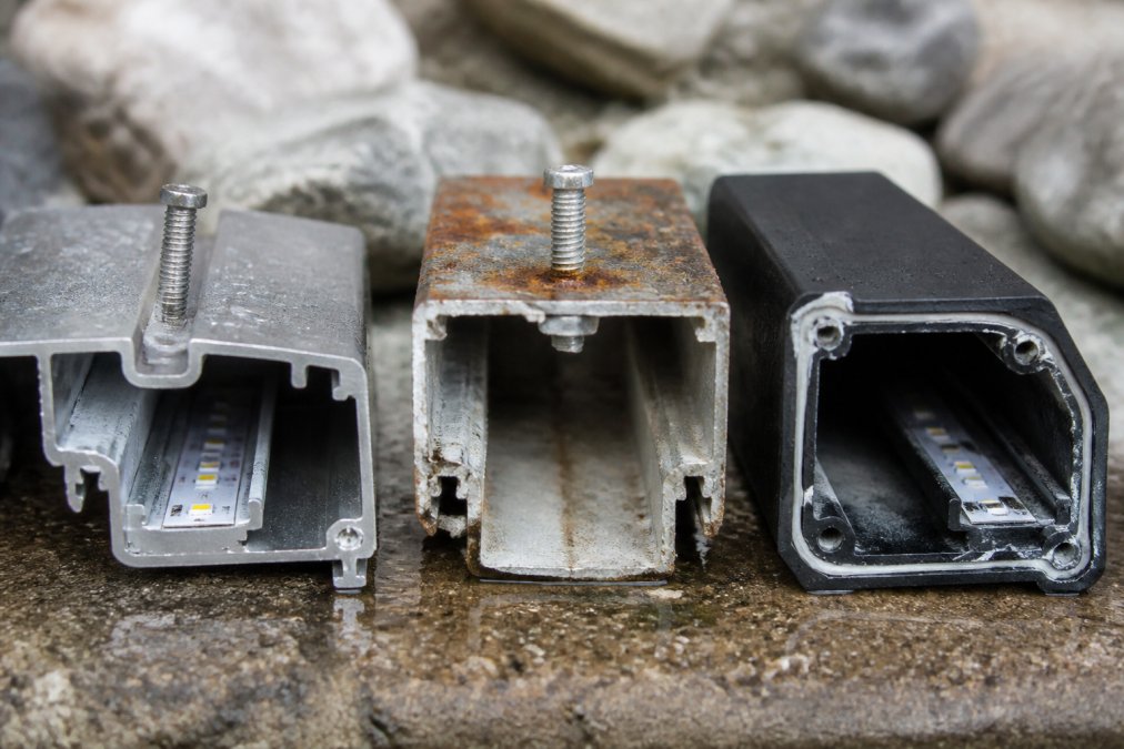 Cross-section comparison of three outdoor smart light housings showing anodized aluminum with stainless fasteners, 304 stainless steel, and UV-stabilized plastic, highlighting corrosion patterns and seal stress points under freeze-thaw cycling