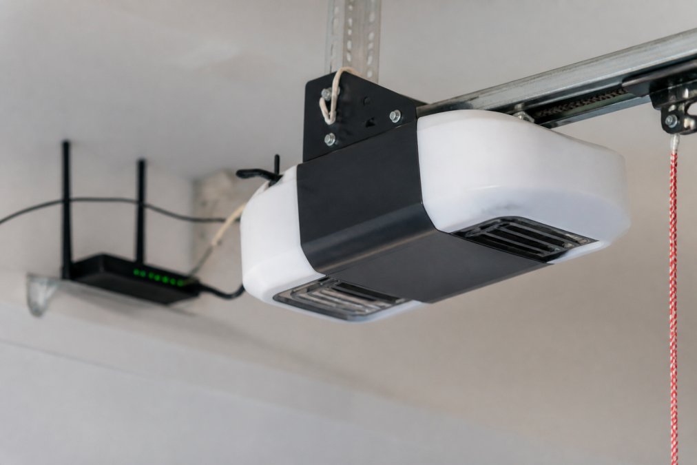 Close-up of a modern smart garage door opener motor mounted on a garage ceiling with WiFi router visible in background, showing the control mechanism and sensor connections