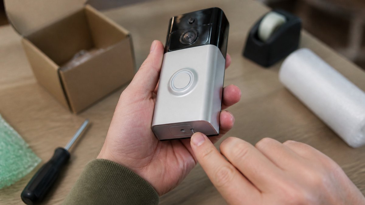 Person's hands holding a smart home doorbell camera with index finger positioned on the small reset button located on the device's base, demonstrating the physical reset action in natural indoor lighting