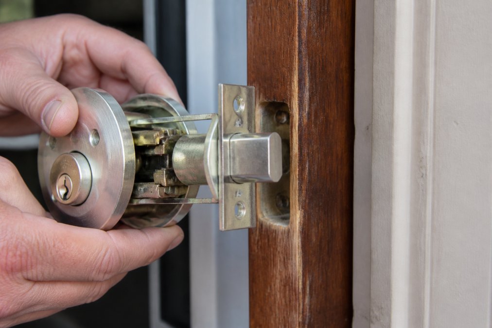 Hands demonstrating the internal mechanism of a residential deadbolt lock showing visible hardened steel construction and bolt throw against a wooden door frame in natural daylight