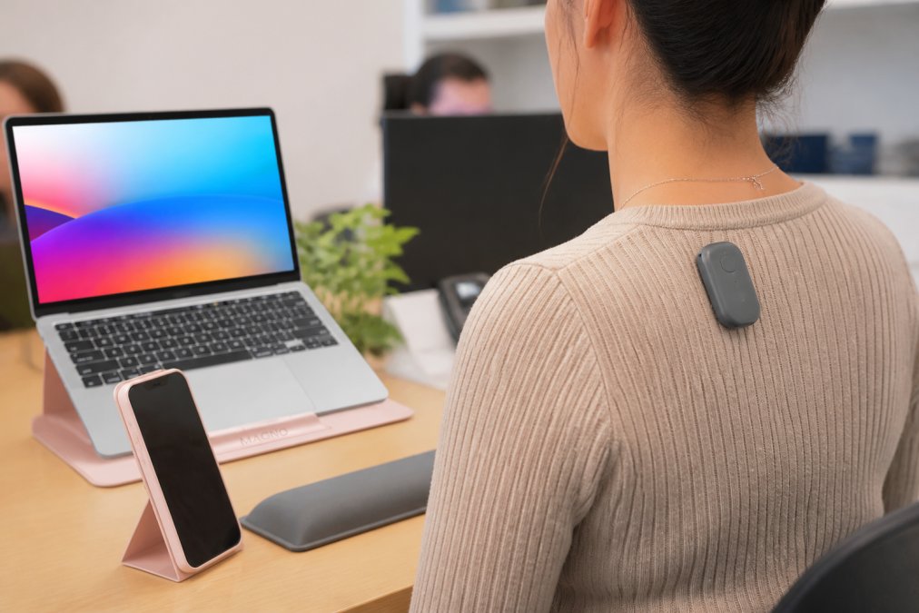 Close-up of a small wearable posture sensor clipped to the upper back of a person sitting at a desk, with a smartphone displaying a posture tracking app nearby, natural office lighting