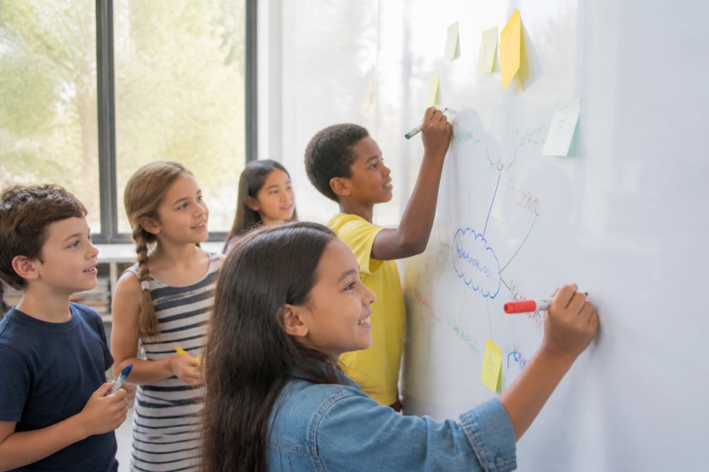 Primary school classroom with students writing on a white magnetic wall surface using dry-erase markers, with children collaborating on a collaborative lesson plan, natural light from windows illuminating engaged faces and colourful marker work on the wall