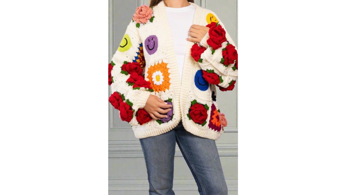 A stylish woman in an oversized chunky knit cardigan with colourful patchwork and smiley faces, standing confidently in a bright, minimal living room with natural light streaming in