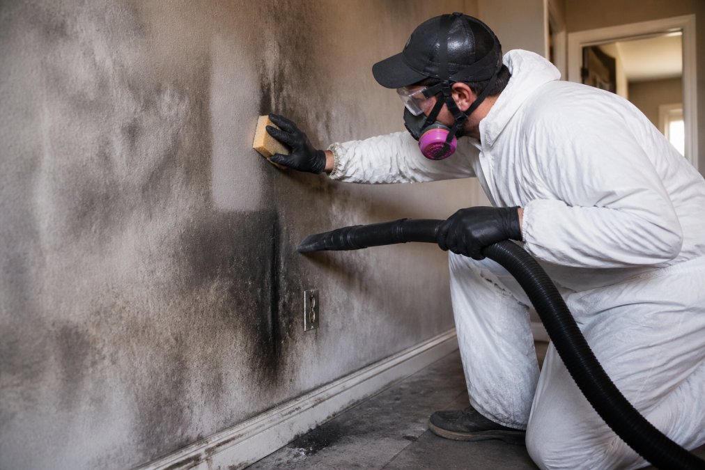 Professional restoration technician in protective gear using HEPA vacuum and dry chemical sponge to carefully clean smoke and soot damage from interior wall surfaces in a residential space