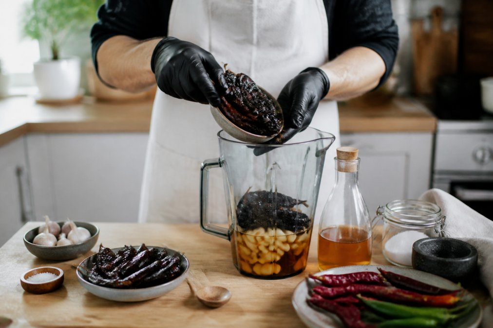 Home cook wearing gloves adding charred chillies to a blender to make smoky hot sauce with vinegar and garlic