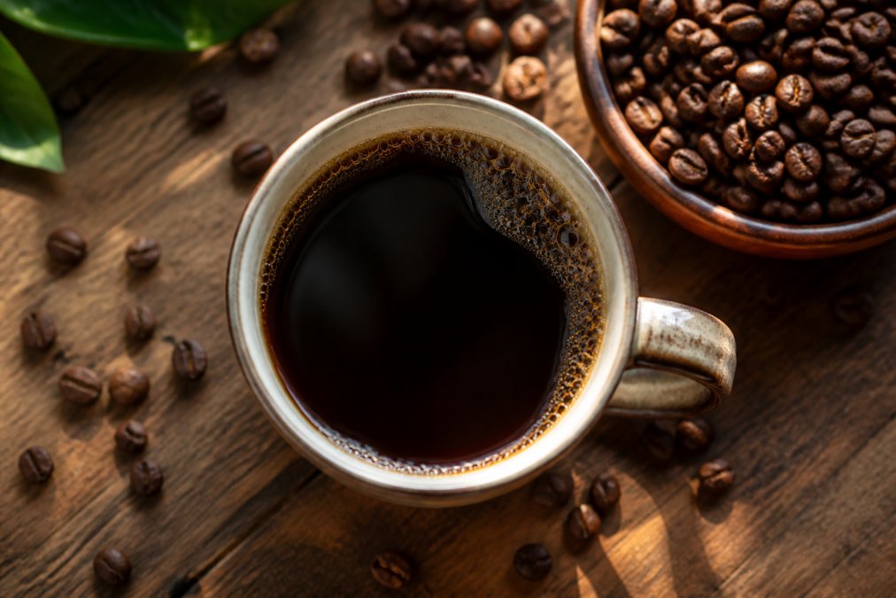 A close-up overhead shot of a freshly brewed cup of black coffee in a ceramic mug on a wooden table, with scattered whole coffee beans and a small bowl of roasted beans nearby, warm natural light